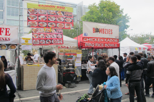 Menschen stehen auf einer Straße außerhalb der Stadt, mit einem Mann in einem grauen und weißen T-Shirt, der eine Wasserflasche hält, und einer Frau in einer blauen Jacke, die einen Kinderwagen schiebt. Im Hintergrund sind Stände mit weißen und roten Zelten und bunten Fahnen zu sehen, sowie Gebäude, Bäume und eine Straßenlaterne.
