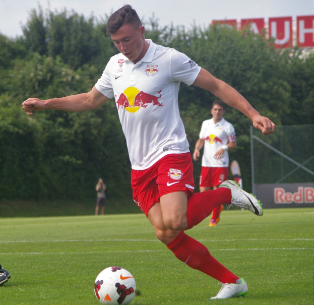 Ein professioneller Fußballspieler tritt einen Ball auf einem grünen Feld mit Bäumen und einem klaren blauen Himmel im Hintergrund, wobei "RB Leipzig" auf einer Tafel hinter ihm zu sehen ist.