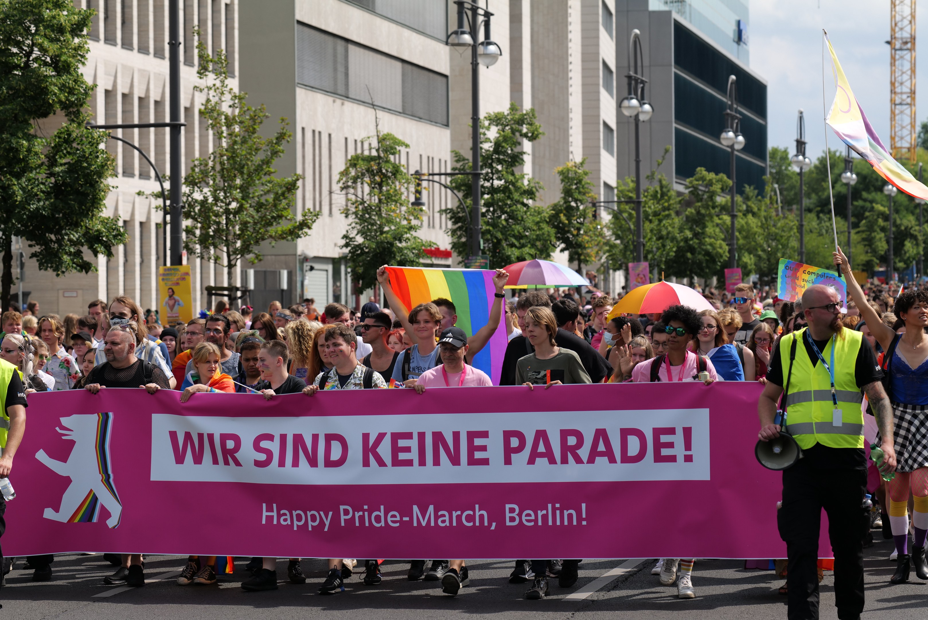 Eine Gruppe von Menschen marschiert bei einer Pride-Veranstaltung in Berlin, hält eine pinkfarbene Fahne mit der Aufschrift "Happy Pride March Berlin" und Schirme und Fahnen, vor einem Hintergrund von Gebäuden, Bäumen, Laternenmasten und einem Turm unter einem klaren blauen Himmel.