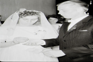 Ein Schwarz-Weiß-Foto einer Braut in einem weißen Kleid und Schleier und eines Bräutigams in einem Anzug, die Ringe tauschen, mit ein paar Menschen im Hintergrund.