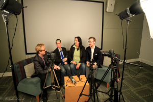 Eine Gruppe von drei Menschen sitzt auf einem Sofa und gibt ein Interview, wobei einer ein Mikrofon hält, ein Aufnahmegerät auf dem Tisch in der Mitte steht, Kameras auf beiden Seiten und ein Projektionsschirm dahinter zu sehen ist.