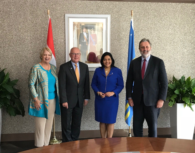Eine Gruppe von Menschen, die um einen Tisch mit Papieren stehen und lächeln, umgeben von Fahnen, Topfpflanzen und einem Foto auf der Wand.