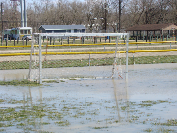 Ein Fußballtor steht in einem überfluteten Feld mit umgebender Wiese und Wasser, mit Hütten, Pfählen, Bäumen, Fahrzeugen und einem klaren blauen Himmel im Hintergrund.