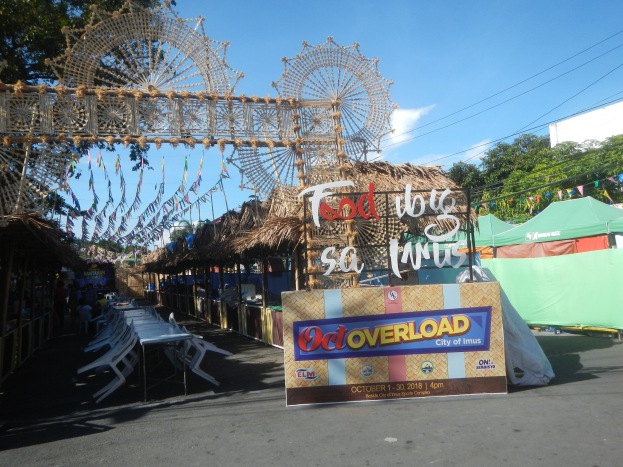 Eine KarnevalsSzene mit einem großen Riesenrad in der Mitte, umgeben von verschiedenen Hütten, Tischen, Stühlen, einem Schild, Dekorationen, Bäumen und Überlandleitungen unter einem bewölkten Himmel.