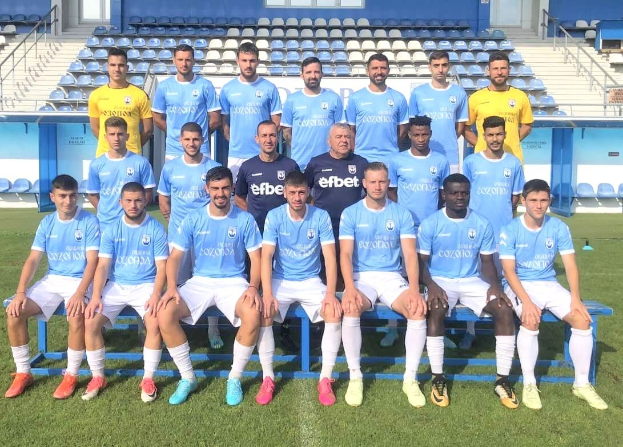 Gruppe von Männern auf einem Fußballfeld, einige sitzen auf Bänken und andere stehen auf dem Rasen, mit Tribünen und anderen Gebäuden im Hintergrund.