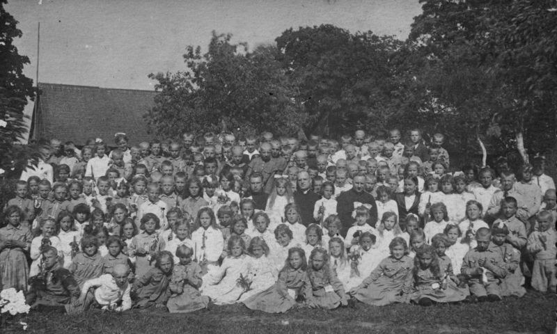 Schwarz-weiß-Foto einer Gruppe von Kindern, die vor einem Haus mit Bäumen und einem klaren Himmel posieren.