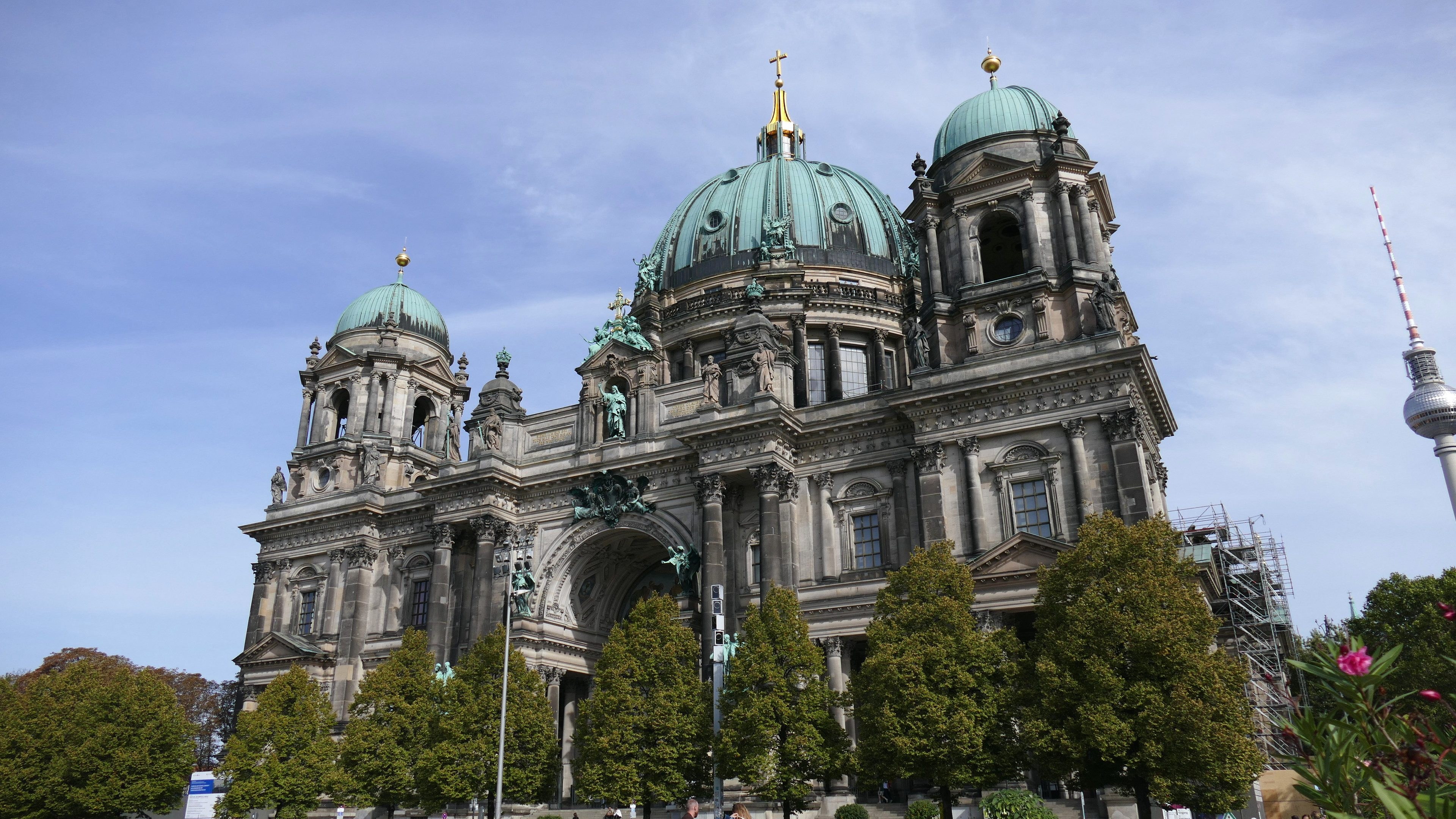 Der Berliner Dom, eine große Kathedrale mit detaillierten architektonischen Elementen wie Fenstern, Säulen, Bögen und Statuen, steht prominent mit Menschen davor, umgeben von Bäumen, Blumenpflanzen und einem Turm unter einem bewölkten Himmel.