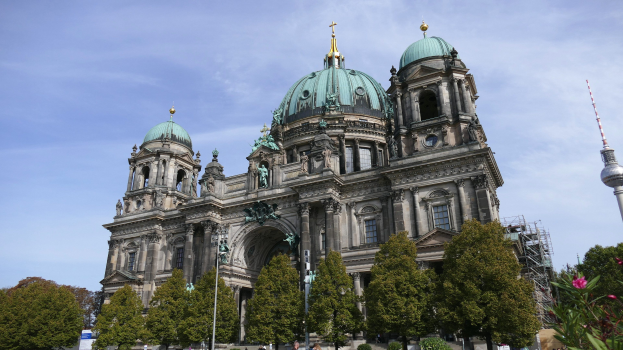 Der Berliner Dom, eine große Kathedrale mit detaillierten architektonischen Elementen wie Fenstern, Säulen, Bögen und Statuen, steht prominent mit Menschen davor, umgeben von Bäumen, Blumenpflanzen und einem Turm unter einem bewölkten Himmel.