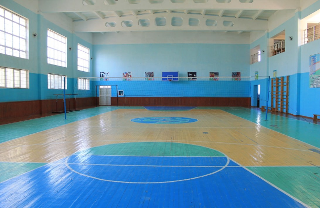 Großer Turnsaal mit blau-weißem Boden, einem Basketballfeld, einem Netz mit Stangen, einer Tür, Fenstern, Wandplakaten, einem Geländer und einem Dach.