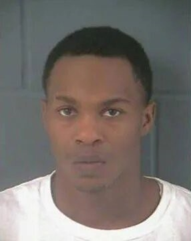 Schwarz-weißes Mugshot eines Mannes in einem weißen T-Shirt, der in die Kamera schaut, mit einer neutralen Wand im Hintergrund.