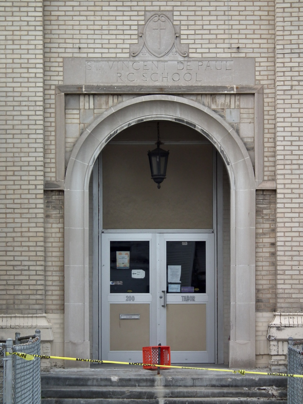 Eingang zu einem Backsteingebäude mit gelbem Absperrband, Treppe mit Maschendrahtzaun, Tür mit Papieren, eine hängende Lampe und eine Schulname an der Wand.