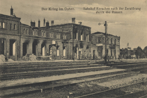 Ein altes Schwarz-Weiß-Foto eines Bahnhofs in Korschen, Deutschland, mit Bahnschienen, Gebäuden, Pfählen, Menschen, Bäumen und Himmel im Hintergrund, mit Text oben und unten, der "Bahnhof Korschen in Ostern, Deutschland" lautet.