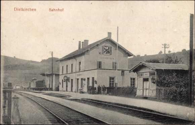 Altes Schwarz-Weiß-Foto eines Bahnhofs in Dielkirchen, Deutschland, mit einem Zug auf den Gleisen, umgeben von Häusern, Strommasten, Bäumen und Menschen, vor einem Hügel- und klaren Himmel.