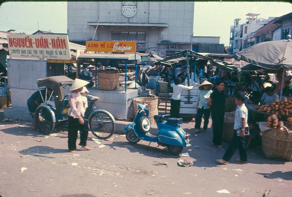 Eine belebte Stadtstraße mit Geschäften, Zelten, Schirmen, einem Auto-Rikscha, einem Motorrad, Passanten und Gebäuden sowie verstreuten Papiermüll in der Vordergrund.