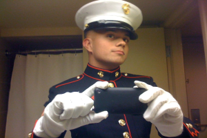 Ein Mann in Uniform steht mit einem Mobiltelefon in der Hand, möglicherweise Fotos machend, vor einer weißen Tür und einem Vorhang, einer weißen Wand rechts und einer Decke oben.