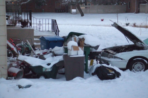 Eine Außenansicht mit Müll in der Mitte, einem Auto auf der rechten Seite, Treppen auf der linken Seite, Bäumen oben und Eis am Boden.
