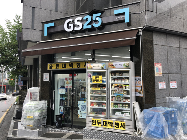 Ein Gebäude mit einem "GS25"-Schild, umgeben von Fahrzeugen, Bäumen und einem klaren blauen Himmel, das einen koreanischen Lebensmittelmarkt mit gekühlten Lebensmitteln im Inneren beherbergt.