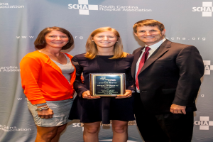 Drei Personen, zwei Frauen und ein Mann, stehen zusammen. Der Mann trägt einen schwarzen Anzug, eine Frau trägt ein orange-schwarzes Kleid und die andere Frau trägt ein schwarzes Kleid und hält einen Preis in der Hand.