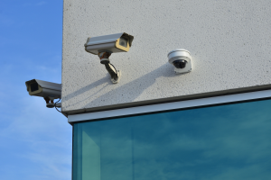 Zwei Sicherheitskameras an der Seite eines Gebäudes montiert, mit einer Glaswand im Vordergrund und einem klaren blauen Himmel im Hintergrund.