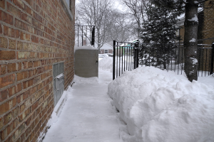 Eine verschneite Szene mit einer Wand auf der linken Seite, Schuppen, Bäumen, Gittern und einem Netz im Hintergrund, alles von Schnee bedeckt, mit Schnee unten.