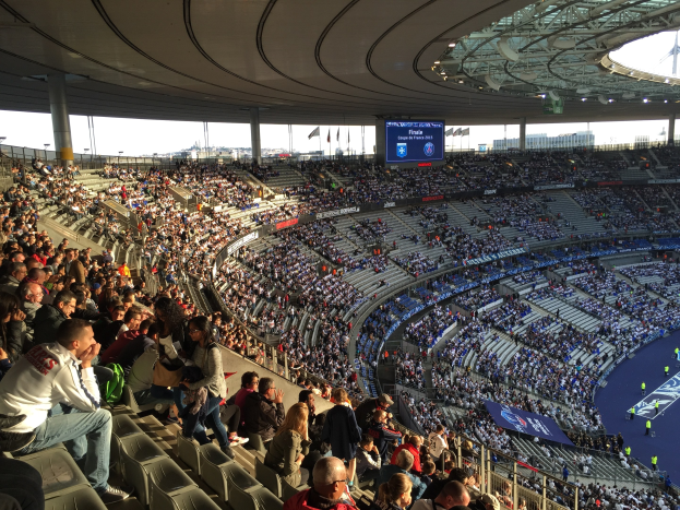 Große Menschenmenge in einem Stadion, das ein Füssballspiel schaut, mit einer Bühne, Fahnen, Masten und einem Bildschirm im Hintergrund, identifiziert als Allianz Arena in München, Deutschland.