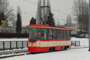 Ein roter und weißer Zug fährt auf schneebedeckten Schienen, umgeben von einem Metallzaun, einem Pfahl, Bäumen, Autos, Gebäuden mit Fenstern, Schildern, Strommasten mit Drähten und einem bewölkten Himmel.