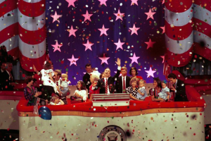 Eine Gruppe von Menschen auf einer Bühne mit einem Rednerpult, das ein Logo, Luftballons und Konfetti trägt, vor einem Hintergrund mit Sternen und einem Vorhang.