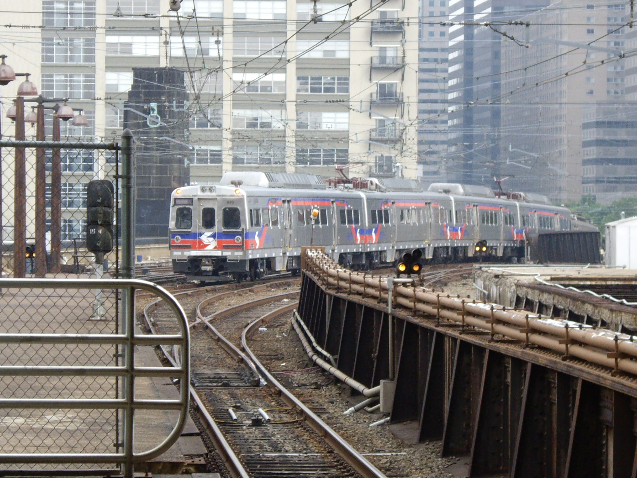 Nahverkehrszug fährt auf Schienen neben hohen Gebäuden mit Bahninfrastruktur und städtischen Elementen im Hintergrund.
