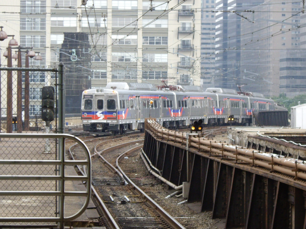 Nahverkehrszug fährt auf Schienen neben hohen Gebäuden mit Bahninfrastruktur und städtischen Elementen im Hintergrund.