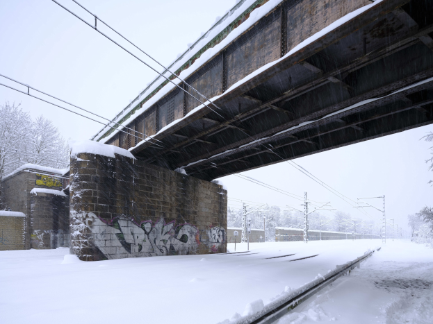 Ein schneebedeckter Bahnübergang unter einer Brücke mit Graffiti, Strommasten, Bäumen und einem Himmel im Hintergrund.