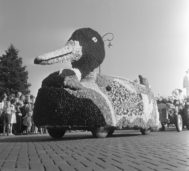 Ein Schwarz-Weiß-Foto eines Entenwagens, der mit Blumen geschmückt ist und von Menschen umringt wird, mit Bäumen und einem klaren Himmel im Hintergrund.