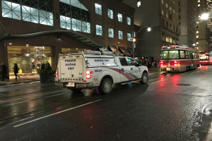 Ein Polizeilaster parkt nachts am Straßenrand einer Stadt, mit Fahrzeugen auf der Straße, Fußgängern auf dem Gehweg, Topfpflanzen, Straßenlaternen, Gebäuden mit Fenstern und einer bewölkten Himmelslandschaft.