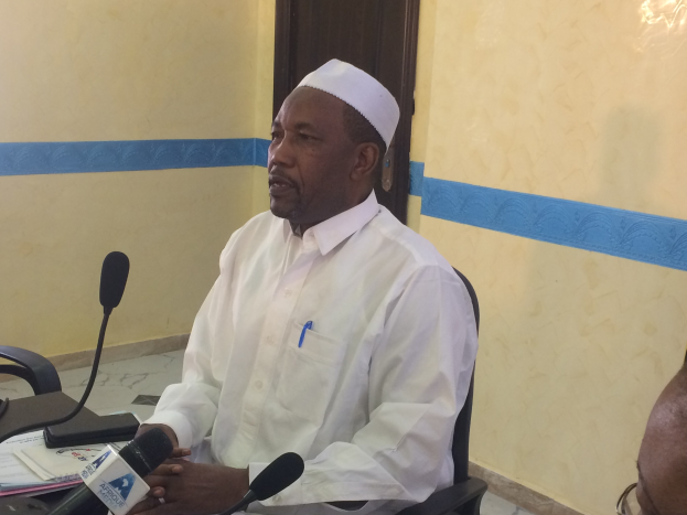 Ein Mann in einem weißen Gewand und Hut sitzt an einem Tisch mit Mikrofonen, spricht, während Papiere und Gegenstände auf dem Tisch liegen, mit einem anderen Menschen mit Brille rechts von ihm, in einem Raum mit einer Wand und einer Tür dahinter.