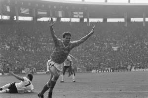 Ein Schwarz-Weiß-Foto eines Fußballspielers, der nach einem Tor jubelt, umgeben von Menschen auf dem Boden, mit Stadiontafeln, Fahnen und Pfosten im Hintergrund unter einem klaren Himmel.