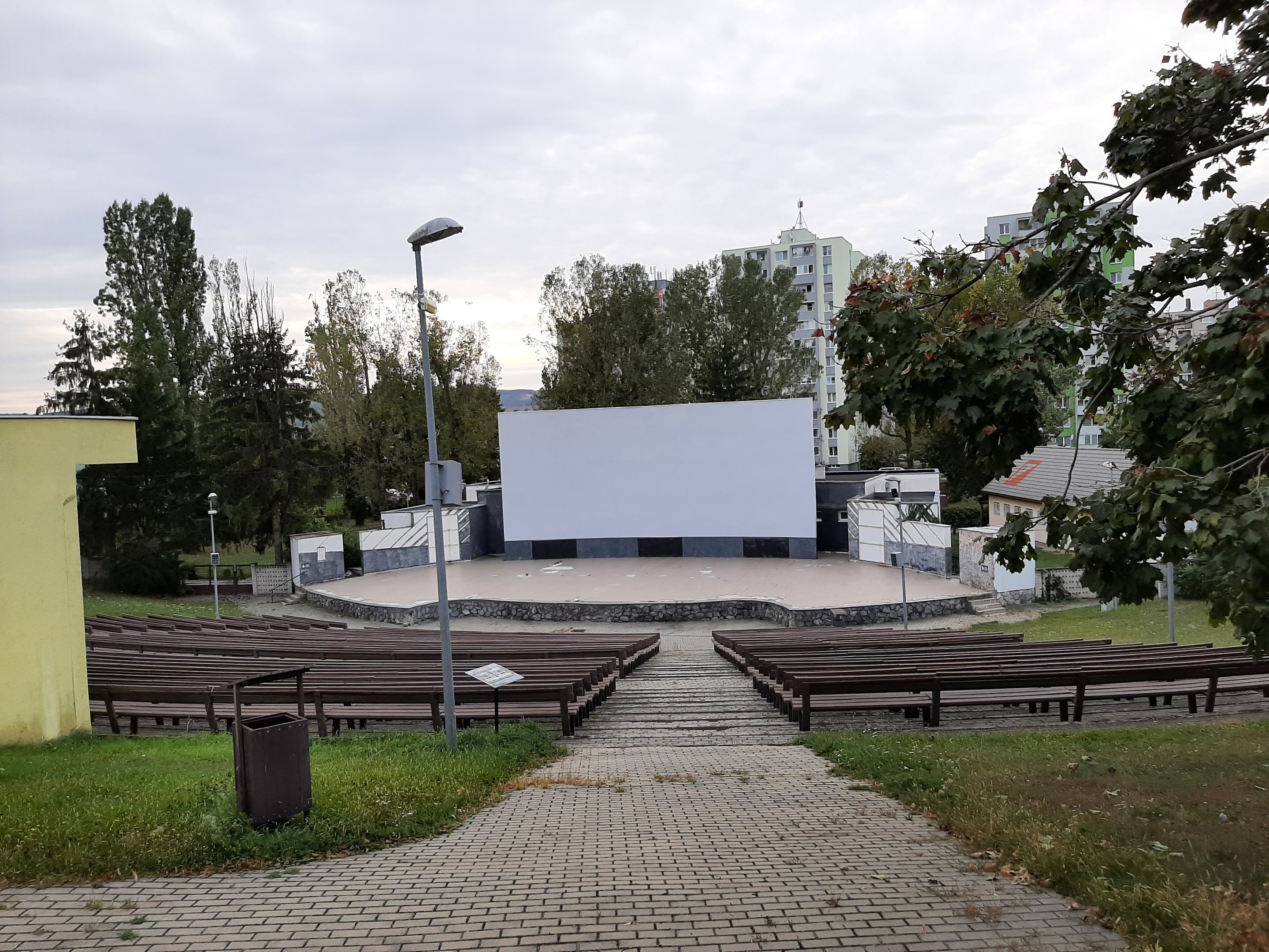 Outdoor movie theater in a park with benches, grass, a trash bin, light poles, trees, and buildings under a cloudy sky.