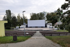 Outdoor movie theater in a park with benches, grass, a trash bin, light poles, trees, and buildings under a cloudy sky.