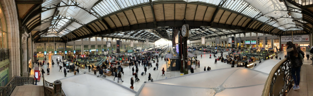 Menschen mit Gepäck durch einen belebten Bahnhof gehen, eine Uhr an der Wand, Anzeigetafeln überkopf und Beleuchtung auf der linken Seite.