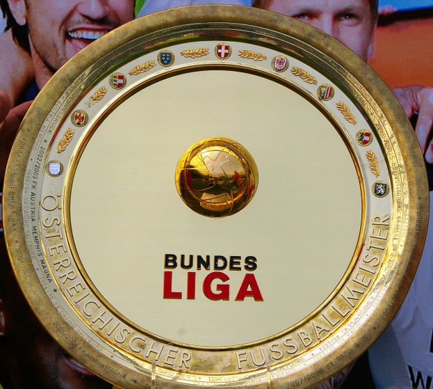 Ein Mann hält einen goldenen Schild mit der Aufschrift "Bundesliga" in die Höhe, im Hintergrund ein Banner mit drei Personen.