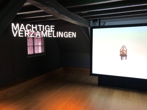 Installationsansicht der Ausstellung 'Machtige Sammlungen' im Museum of Modern Art in Berlin, mit einem großen Bildschirm mit Text an der Wand, einem Fenster links und einem Teil der Decke oben.