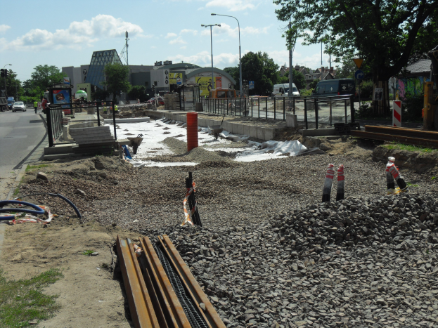Baustelle mit verschiedenen Materialien und Geräten am Straßenrand, Fahrzeuge und Gebäude im Hintergrund.