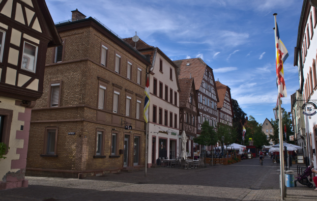 Headsteinstraße in Rothenburg ob der Tauber, Deutschland, gesäumt von Gebäuden mit Fenstern, Bäumen und Fahnenstangen, mit wenigen Menschen und Fahrzeugen auf der Straße unter einem bewölkten Himmel.