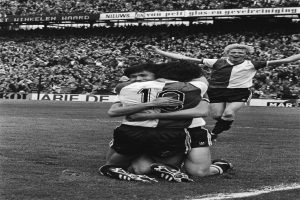 Drei Fussballspieler umarmen sich auf dem Boden und feiern ein Tor vor einer jubelnden Menge in einer schwarz-weissen Stadion-Szene.