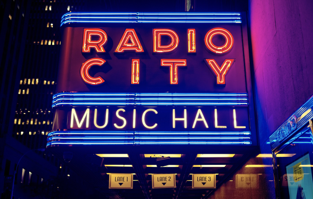 Radio City Music Hall in New York City, hell erleuchtet bei Nacht, umgeben von hohen Gebäuden, mit einer 'Radio City'-Leuchtschrift und zusätzlichen Schildern auf der rechten Seite.