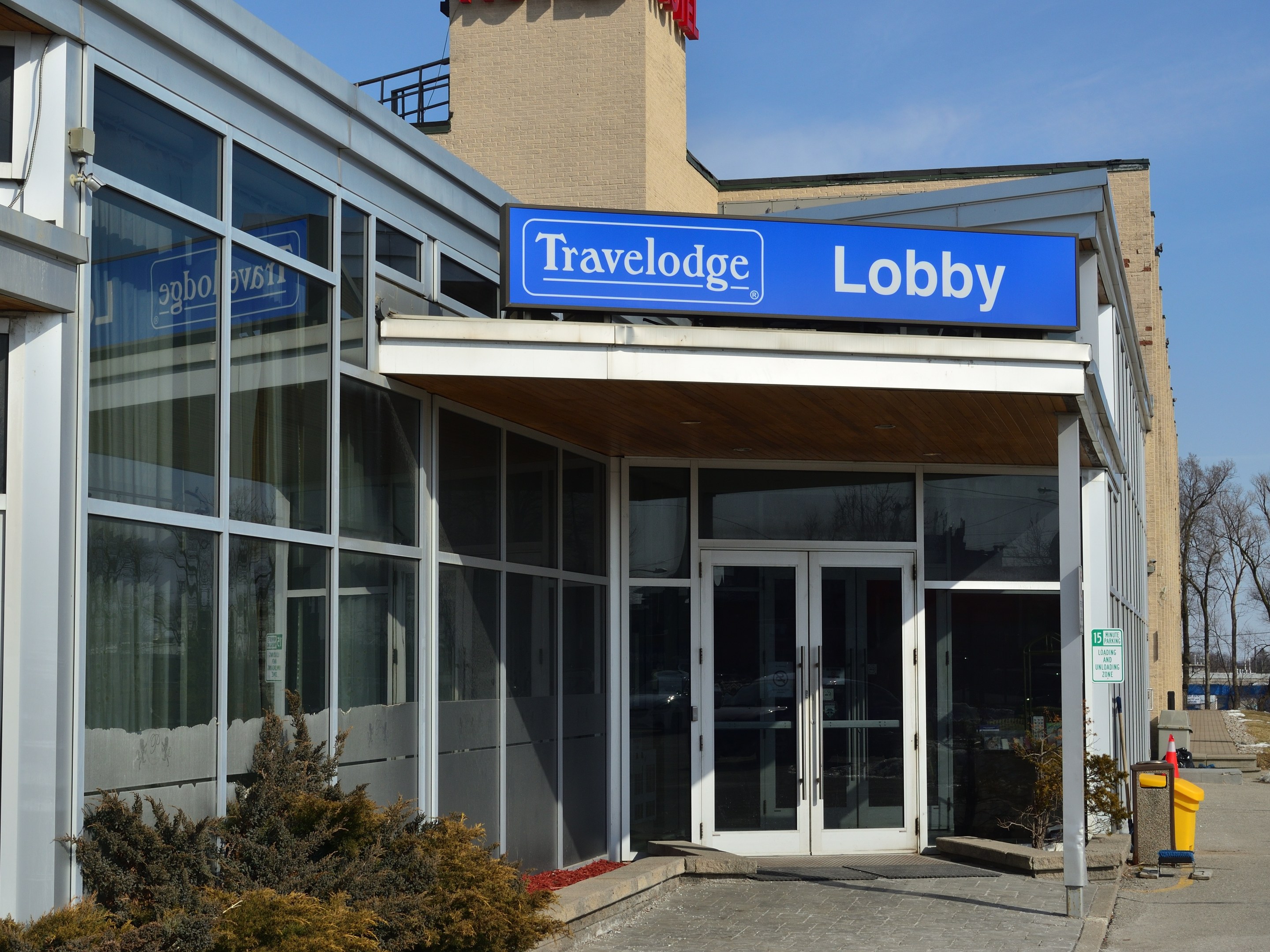 Außenansicht des Travelodge Hotels in Ottawa, Ontario, mit Glaswänden und -türen, einem Schild, Topfpflanzen im Vordergrund, Bäumen und einem klaren blauen Himmel.