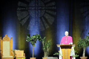 Mann in rosa-weißem Kleid steht neben einem Rednerpult in einem Auditorium, mit Pflanzen und zwei Stühlen daneben und einer bunten Wand im Hintergrund.