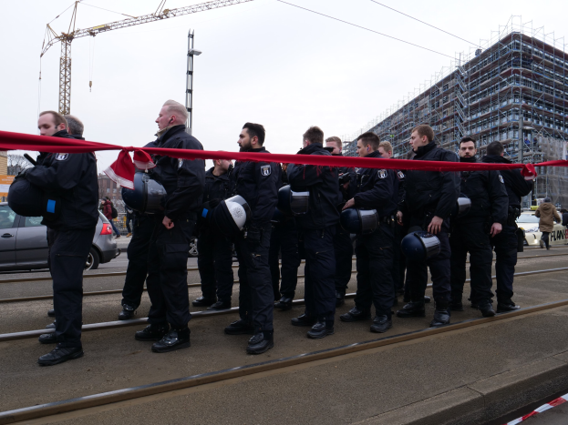Eine Gruppe von Polizisten in schwarzen Uniformen und Helmen steht in einer Reihe auf einem Gleis, mit einem roten Band davor, während im Hintergrund Fahrzeuge, Passanten und Gebäude sowie ein klarer blauer Himmel zu sehen sind.
