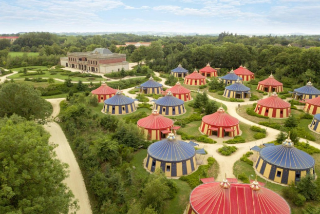 Luftaufnahme eines großen Zirkuszeltes auf einer Wiese umgeben von Bäumen, Pflanzen und Gebäuden unter einem bewölkten Himmel.