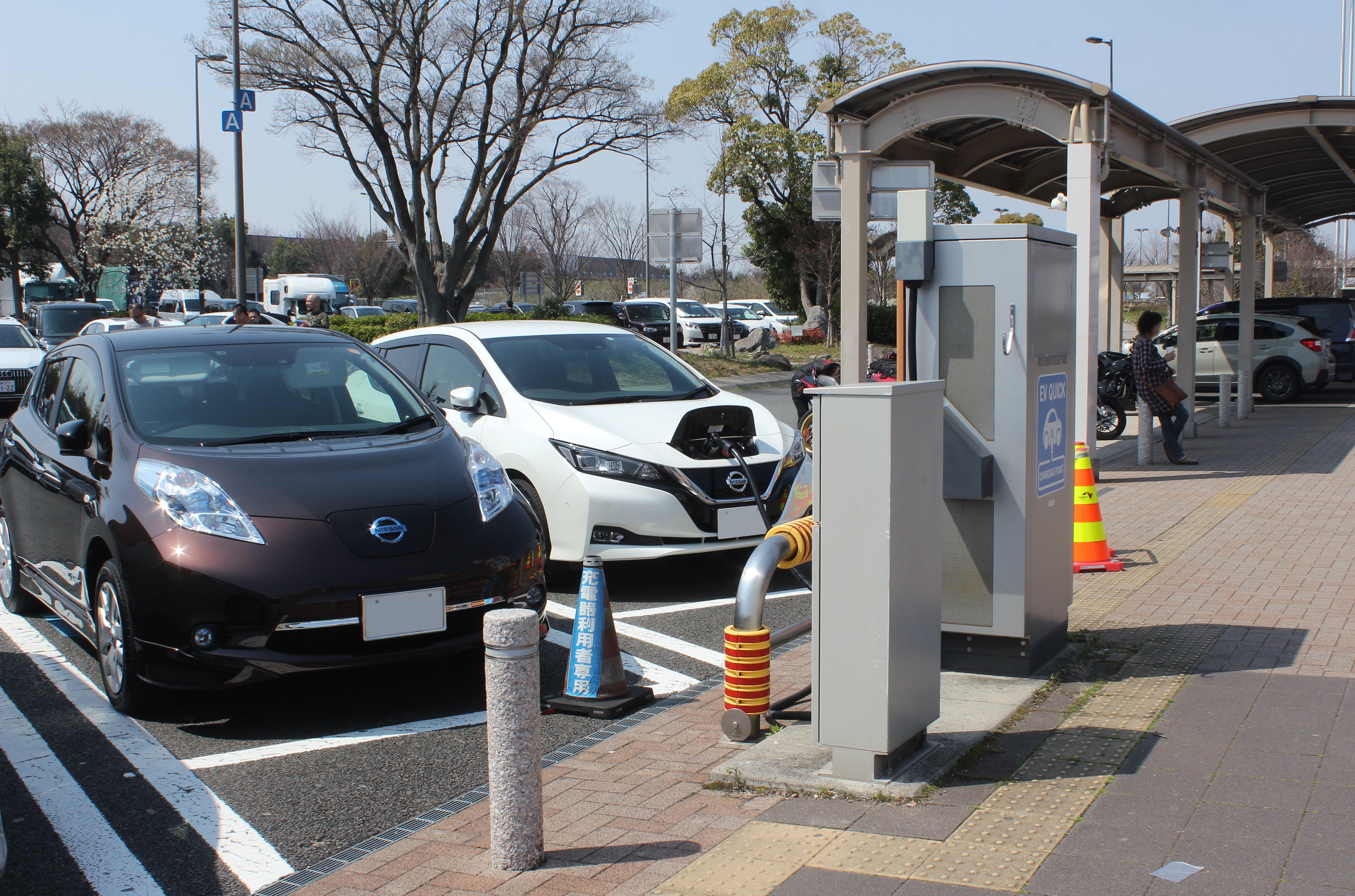 Elektroauto-Ladestation in Japan mit Autos auf der Straße, einer Person auf dem Gehweg, Verkehrskegeln, einem Schuppen, Polen, Lampen, Schildern, Bäumen, Pflanzen und einem Himmel im Hintergrund.