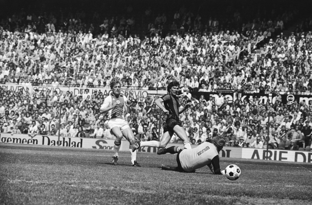 Schwarzes Foto von Männern, die auf einem Feld Fussball spielen, mit Zuschauern im Hintergrund und einer 'Bundesliga 1978-79 Ajax 1-1 Hannover 96'-Fahne.