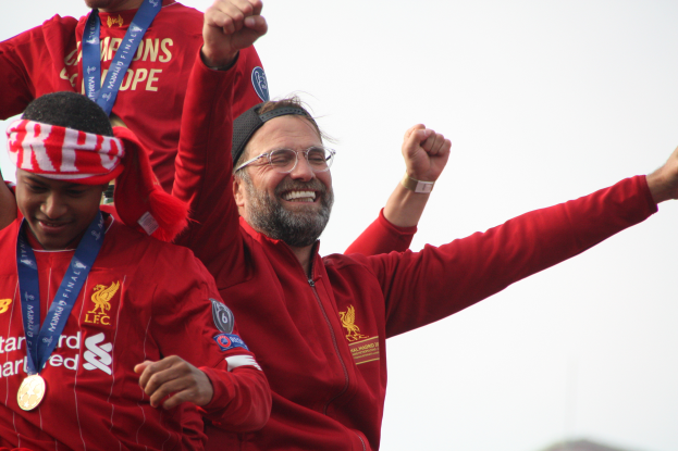 Liverpool-Trainer Jürgen Klopp feiert mit seinen Spielern nach dem Gewinn der UEFA Champions League, während er eine rote Jacke trägt und zwei medalientragende Personen in der Nähe sind und der Himmel im Hintergrund zu sehen ist.